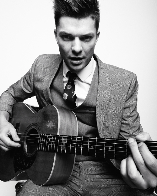 Unique black and white portrait photo with light background of man sitting and posing playing a guitar. 