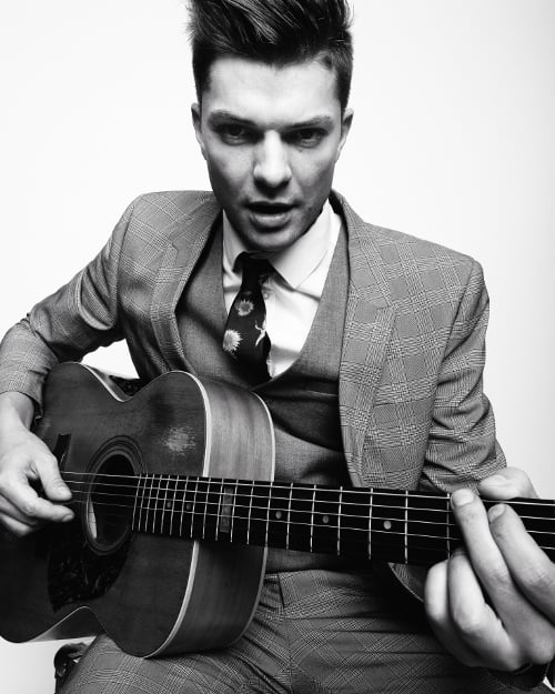 Unique black and white portrait photo with light background of man sitting and posing playing a guitar. 